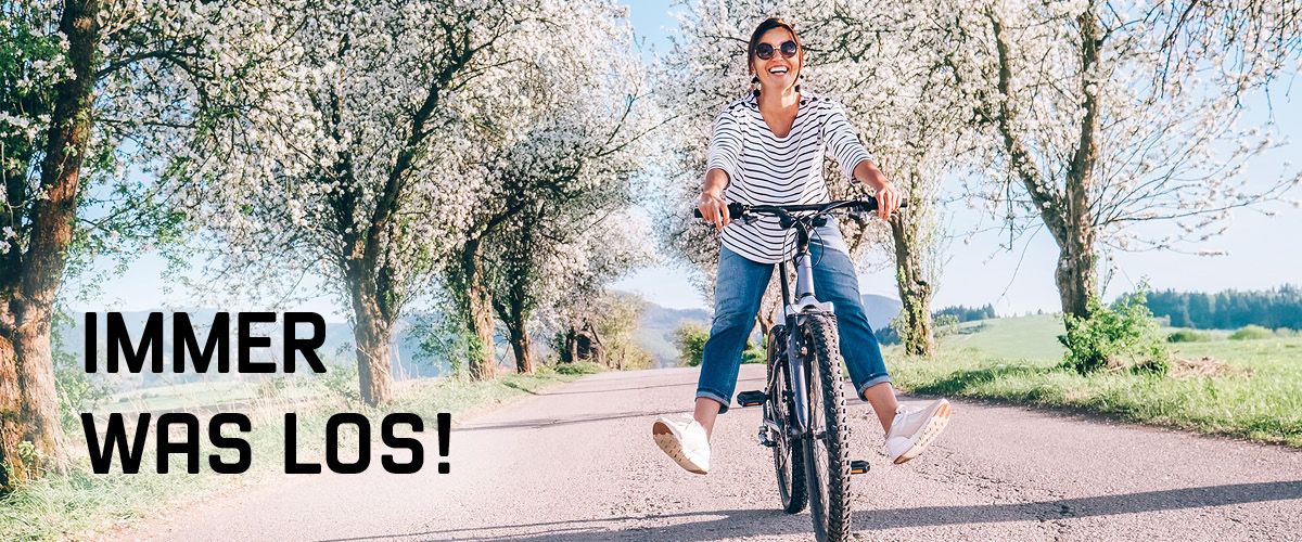 Frau fährt lachend Fahrrad auf einer von blühenden Bäumen gesäumten Straße an einem sonnigen Tag.