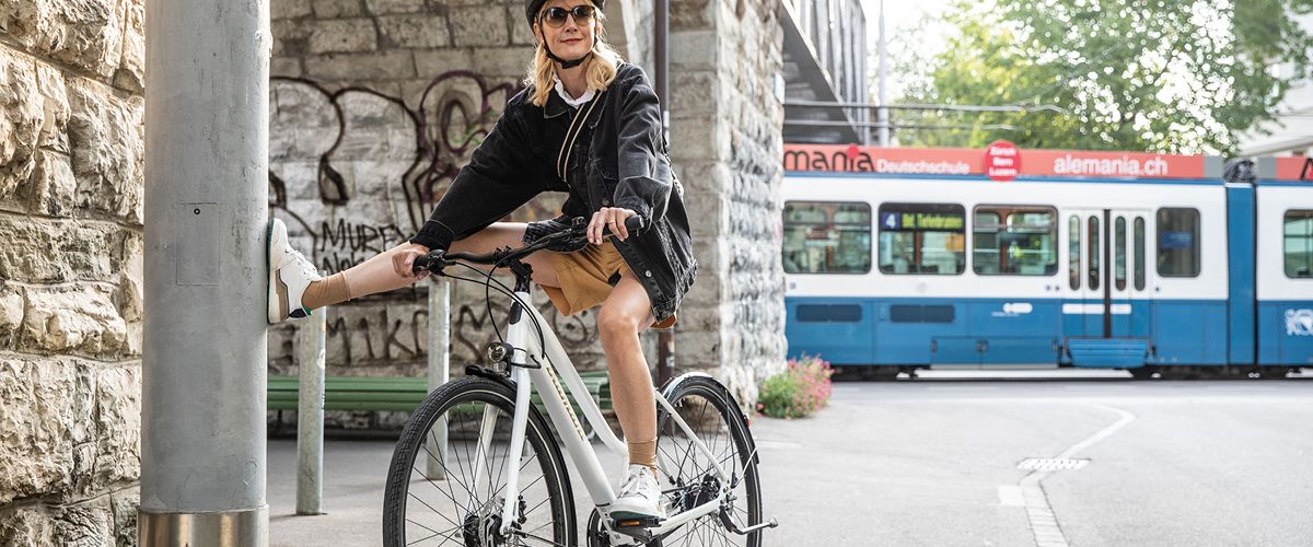 Eine junge Frau auf einem Citybike in der Stadt, im Hintergrund ist eine Straßenbahn und ein Graffiti an einer Wand zu sehen 